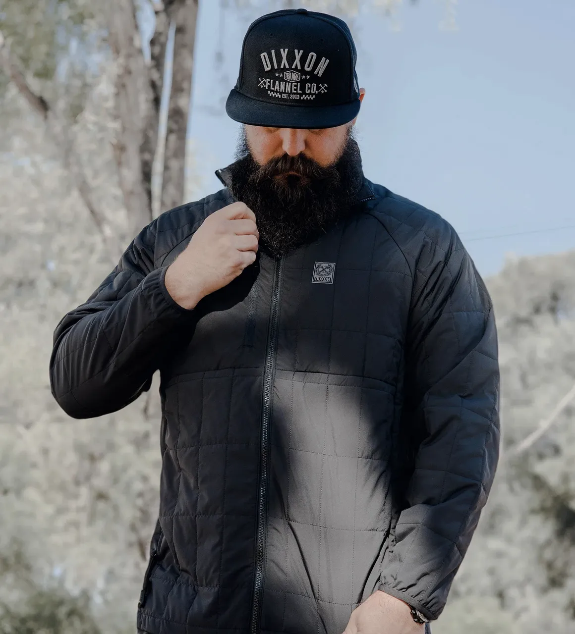 A person with a long, dark beard wearing a black quilted jacket and a black baseball cap with "DIXXON FLANNEL CO." embroidered on the front. They are looking down while adjusting the collar of the jacket.