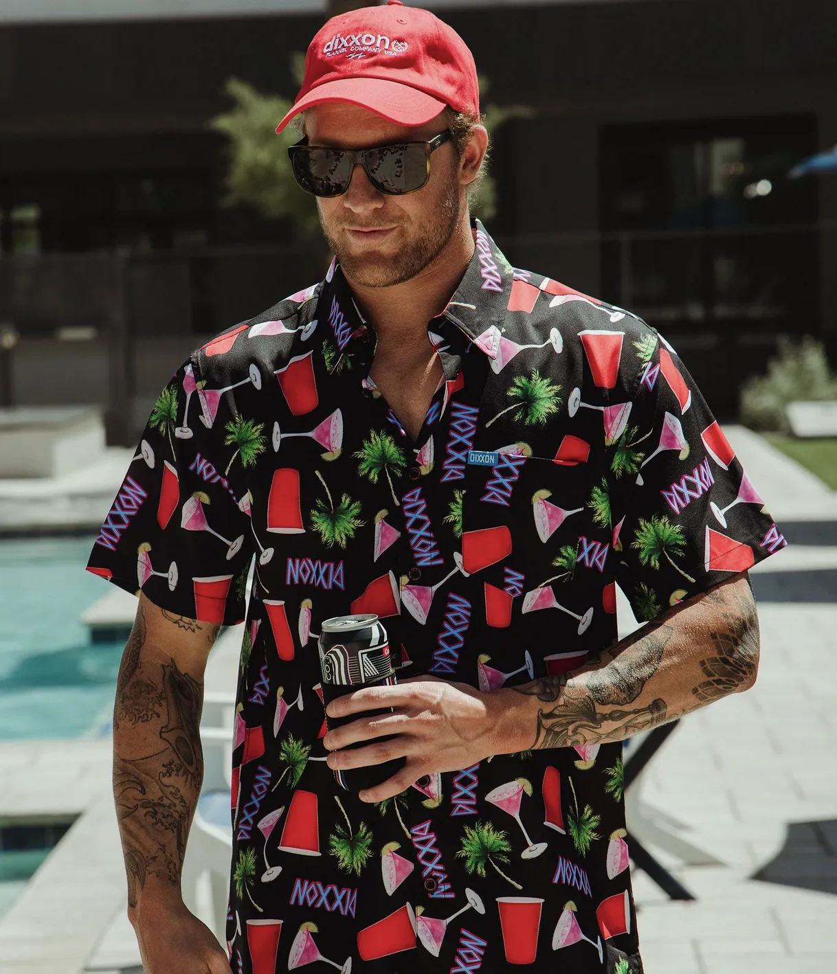 A man standing by a pool wearing a vibrant black Dixxon party shirt with a neon-style pattern of palm trees, red cups, pink cocktails, and the Dixxon logo. He is wearing a red Dixxon ball cap and dark sunglasses while holding a canned beverage.