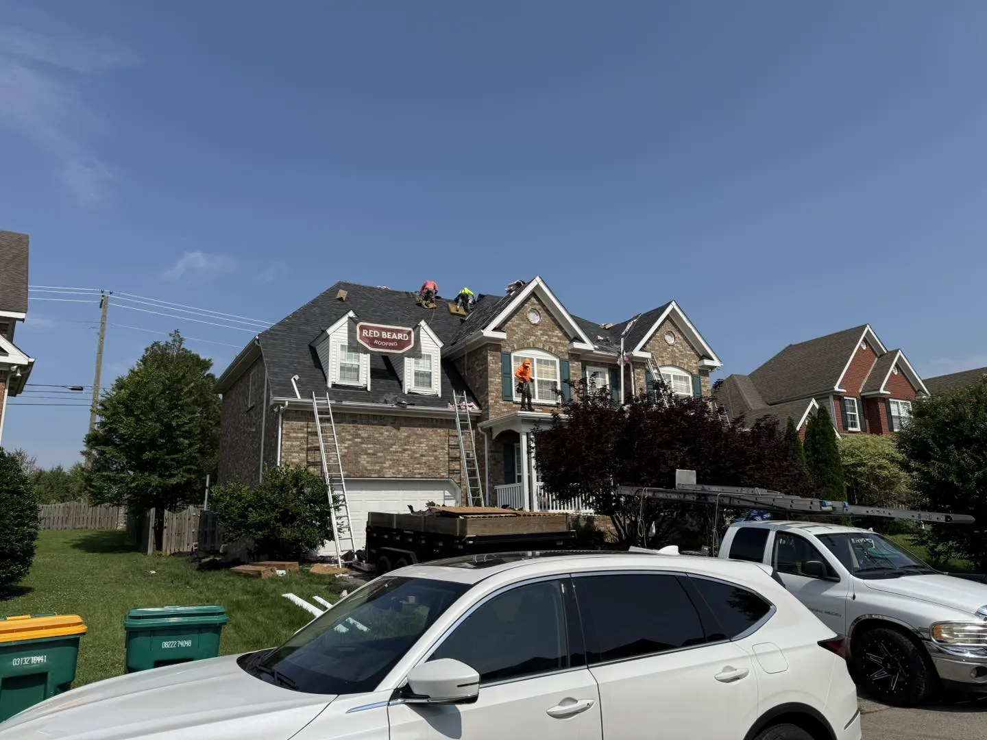 Roof-Replacement underway on a two-story brick residential house by Red Beard Roofing crew, with workers on the roof, ladders set up, and a debris trailer parked in the driveway after Storm Damage