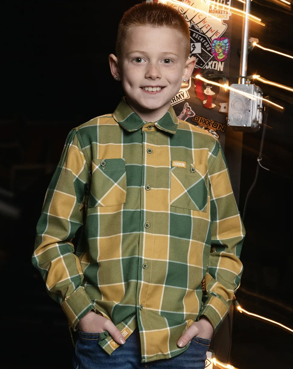 A young boy smiles while wearing a green and gold plaid Dixxon flannel with a custom "DIXXON" logo tag on the chest pocket, standing against a background covered in skate and moto-culture stickers.