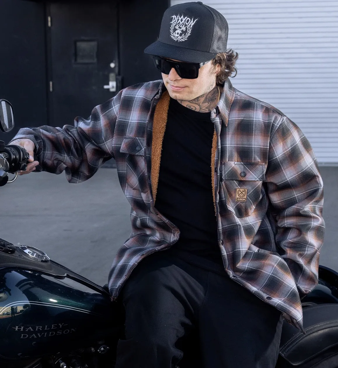 A person wearing a black baseball cap, sunglasses, and a black and orange plaid flannel jacket with a brown sherpa lining. They are seated on a dark green Harley-Davidson motorcycle.