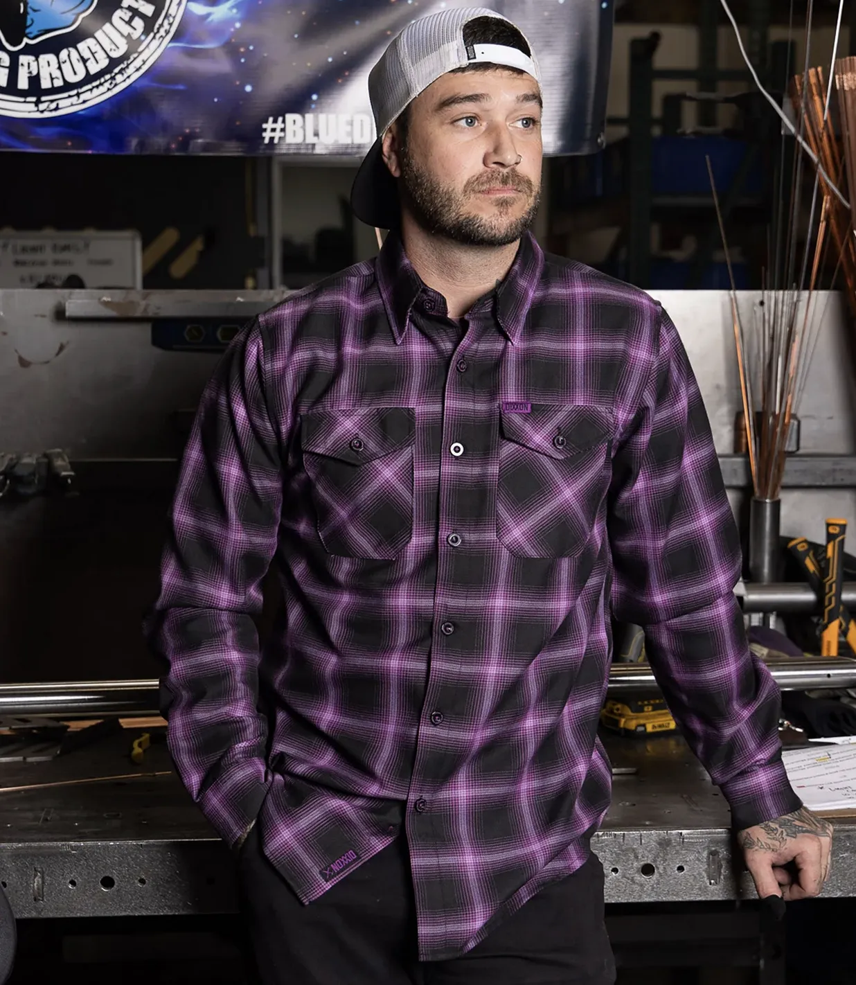 A man with a beard wearing a purple and black plaid Dixxon flannel and a white baseball cap, leaning against a metal workbench in an industrial shop setting.