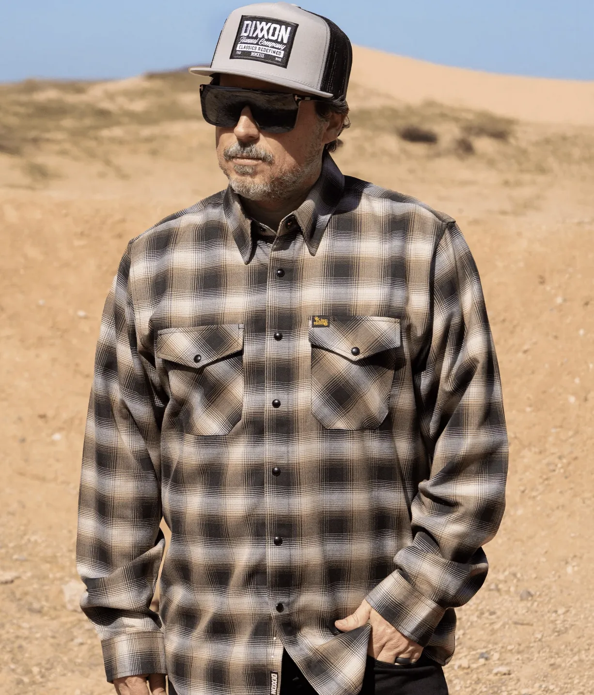 A man wearing a tan, black, and grey plaid Dixxon flannel, dark sunglasses, and a grey and black trucker hat featuring the Dixxon "Classics Redefined" patch. He is standing outdoors in a sandy, desert-like environment.