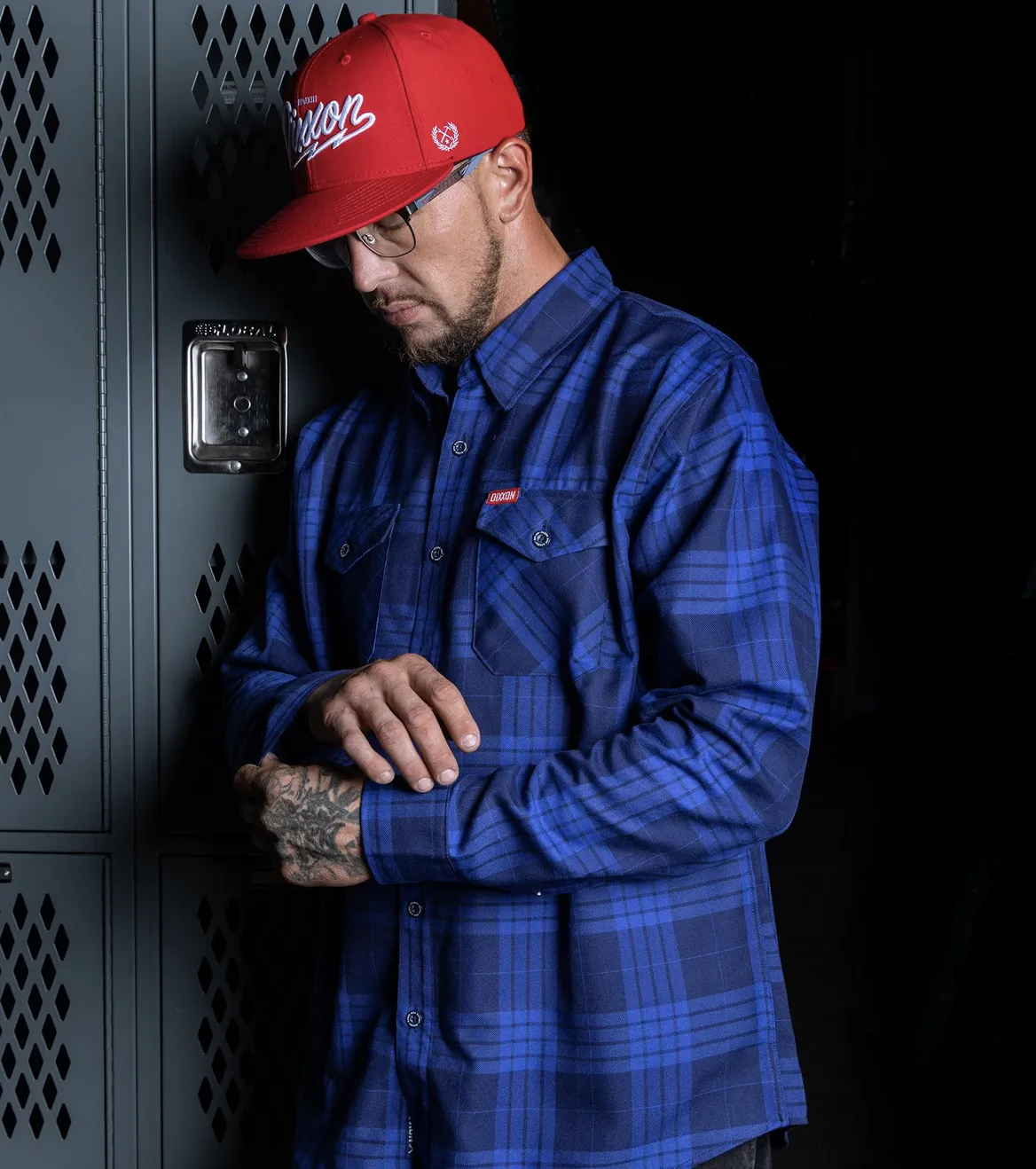 A man with hand tattoos leaning against gray industrial lockers, wearing a blue and navy plaid Dixxon flannel with a red logo tab and a matching red Dixxon cap.