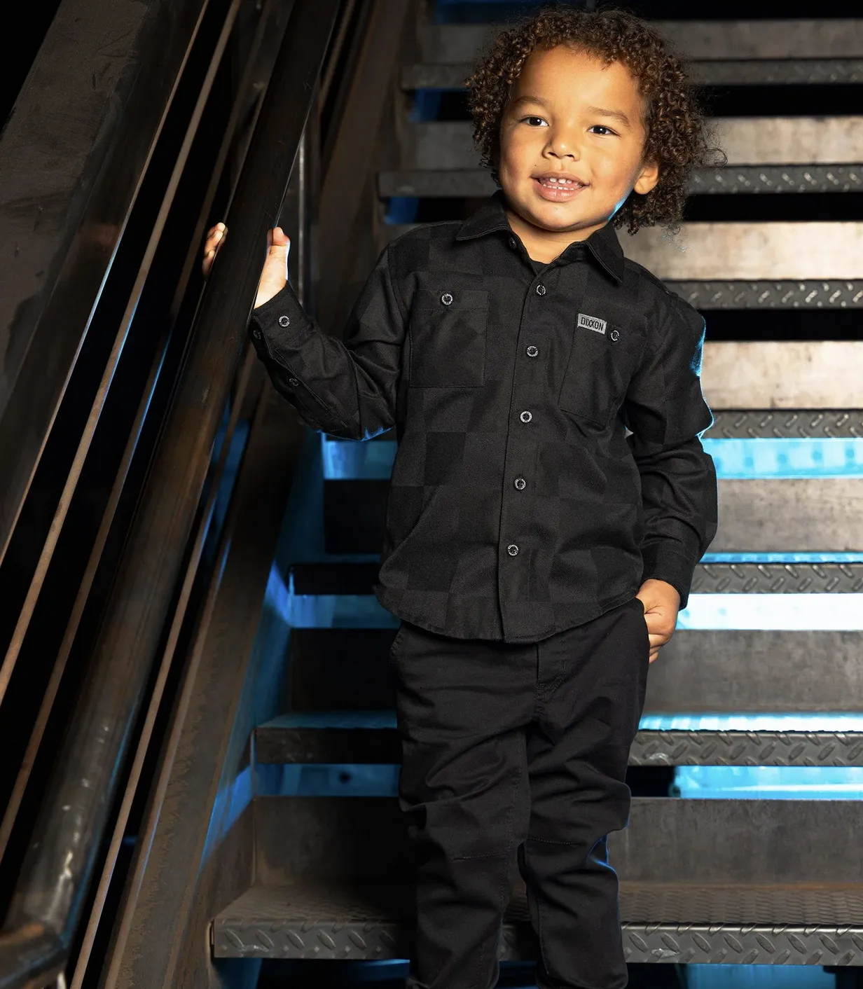 A young child smiling on a metal industrial staircase, wearing a solid black Dixxon youth flannel with signature chest pockets and a custom logo patch.