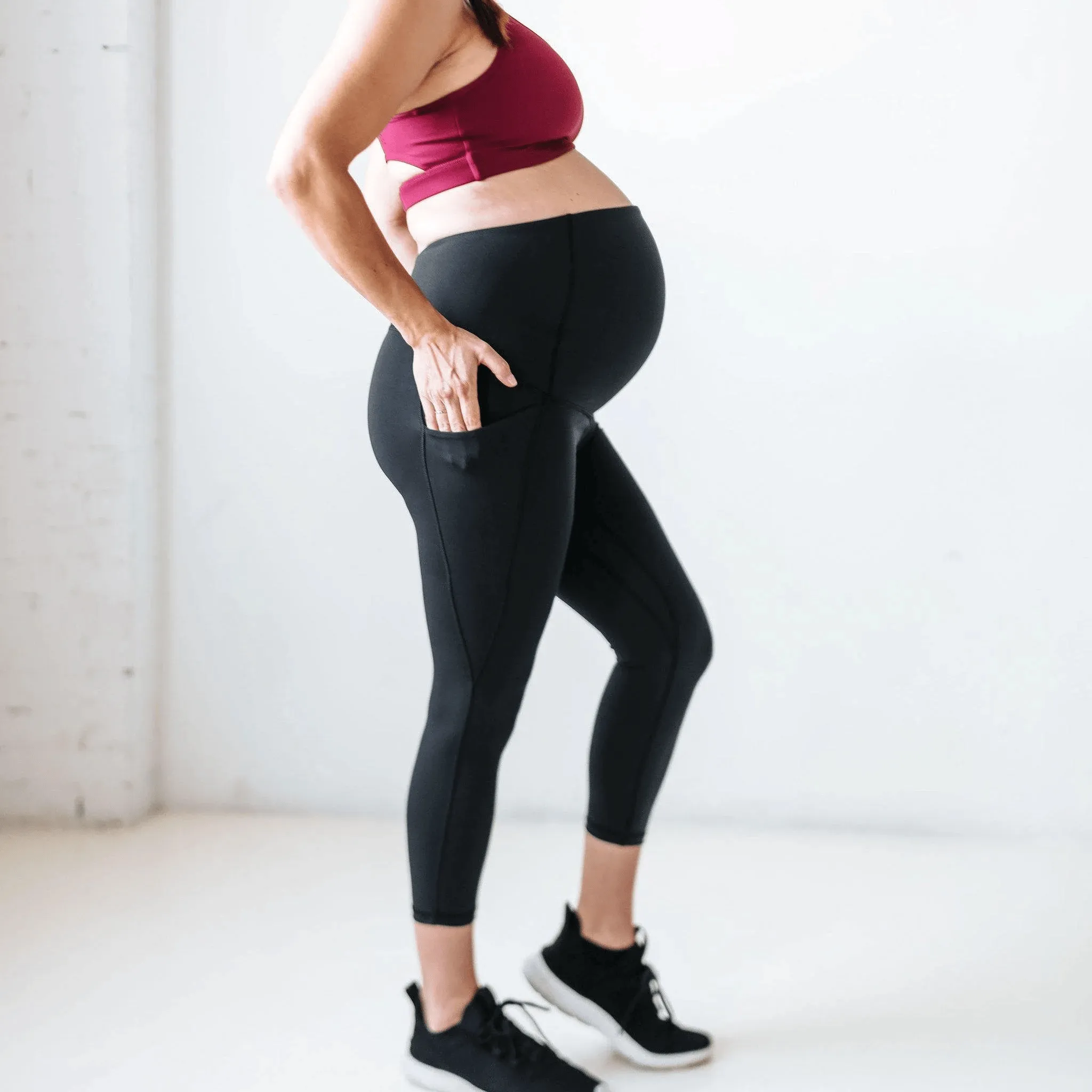 Side view of a pregnant woman wearing black maternity capri leggings and a burgundy sports bra, highlighting the supportive waistband and side pocket.