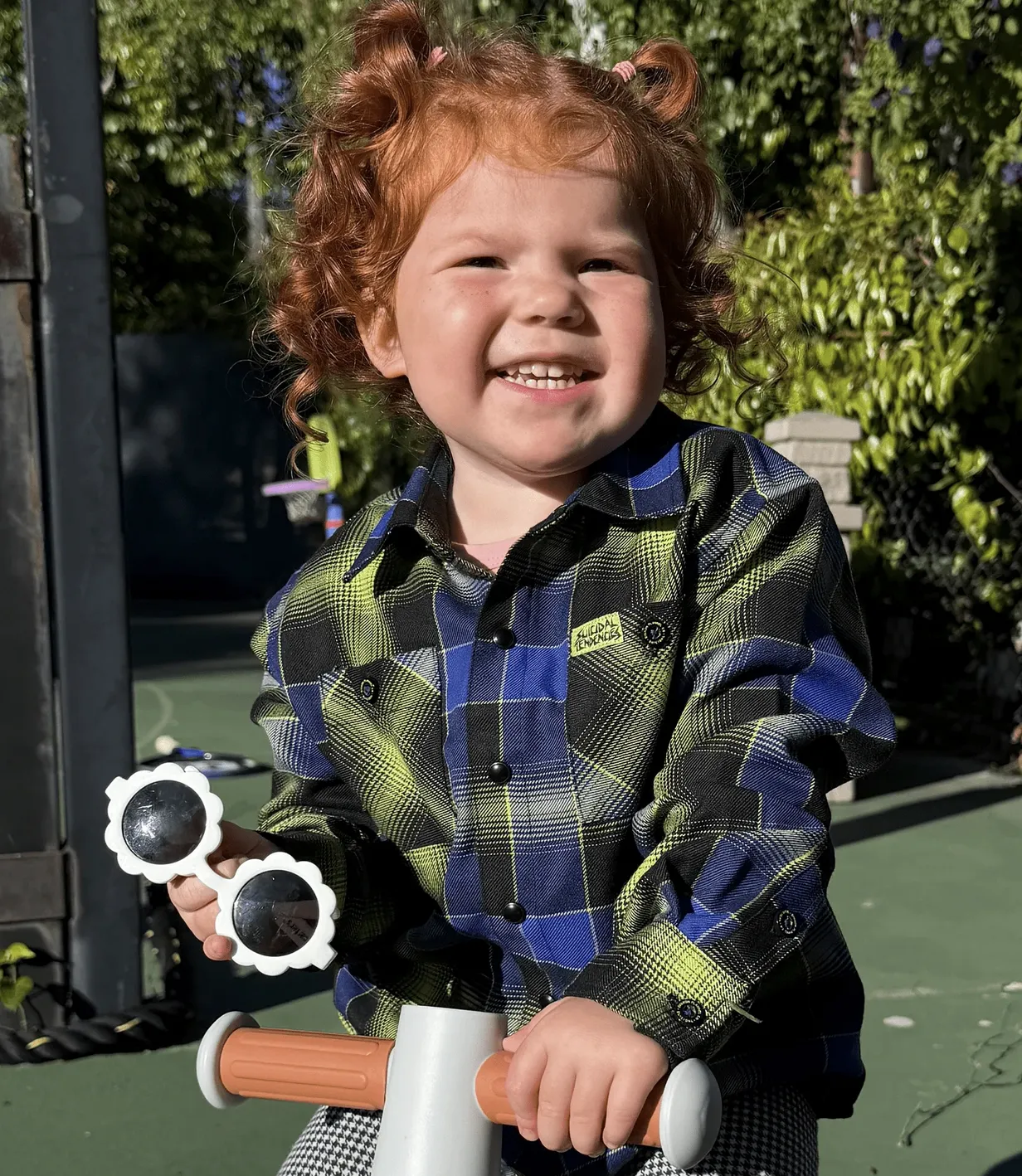 A young child with red hair laughing while riding a toy scooter, wearing a blue and neon green plaid Suicidal Tendencies x Dixxon collaboration flannel.
