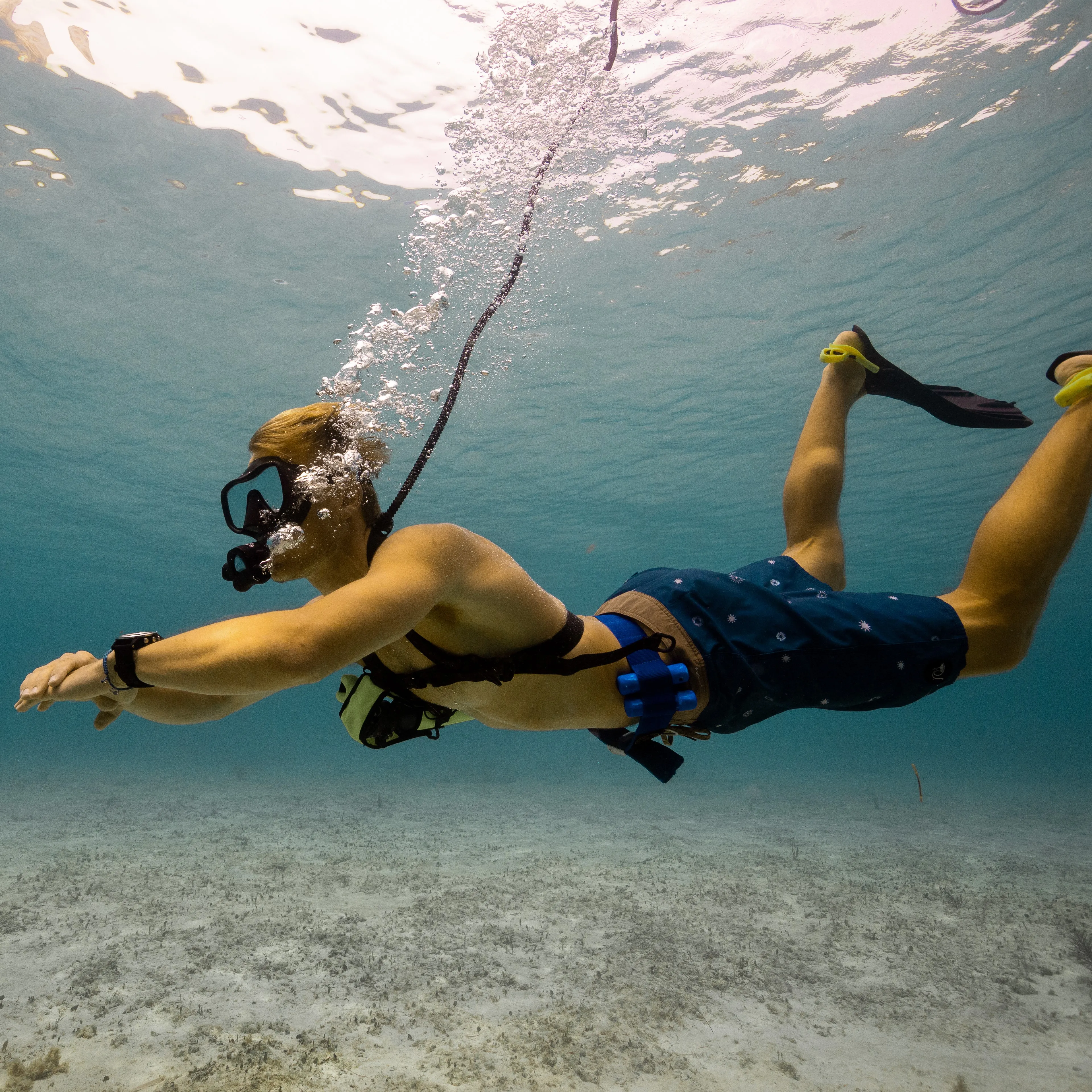 Person diving underwater with a portable tankless dive system, breathing through a hose connected to a floating compressor on the surface.