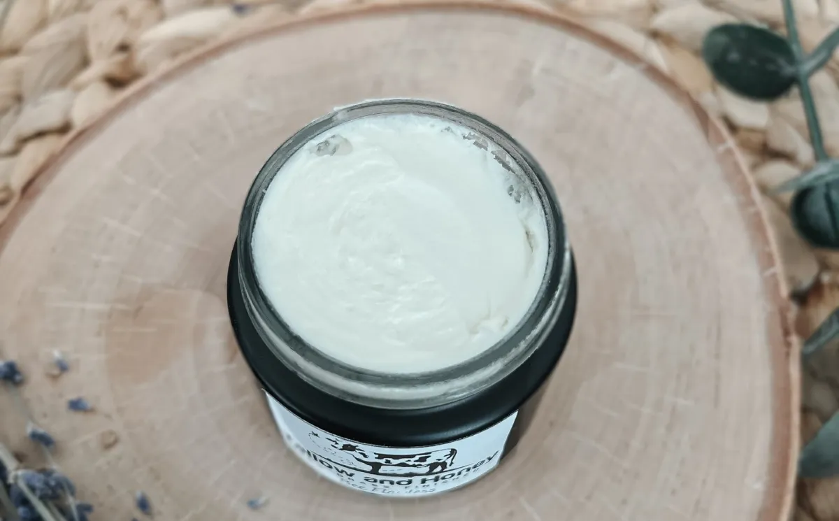 A close-up, overhead shot of a small, dark jar containing a creamy, white tallow and honey balm. The jar sits on a round wooden slice with visible grain, placed on a woven mat. Sprigs of lavender and greenery are visible near the edges, suggesting natural, soothing ingredients.