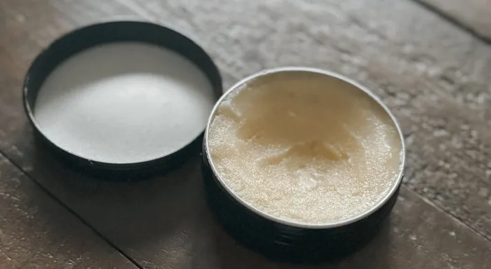 An open metal tin of thick, whipped tallow cream sitting on a rustic wooden surface next to its black lid.