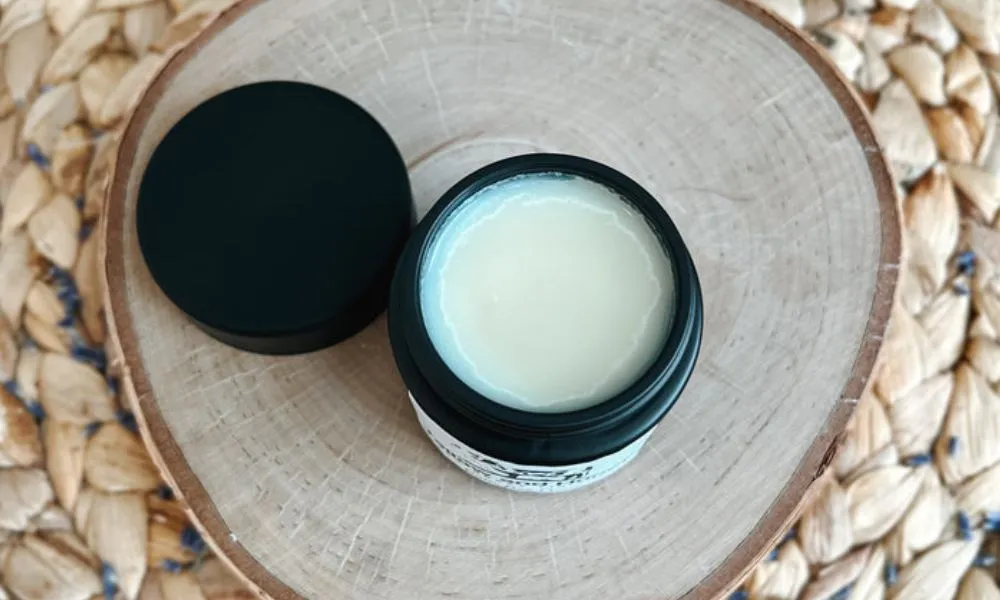 An open jar of smooth, white Beef Tallow balm resting on a rustic wooden coaster, displaying the rich, solid texture of the natural moisturizer.