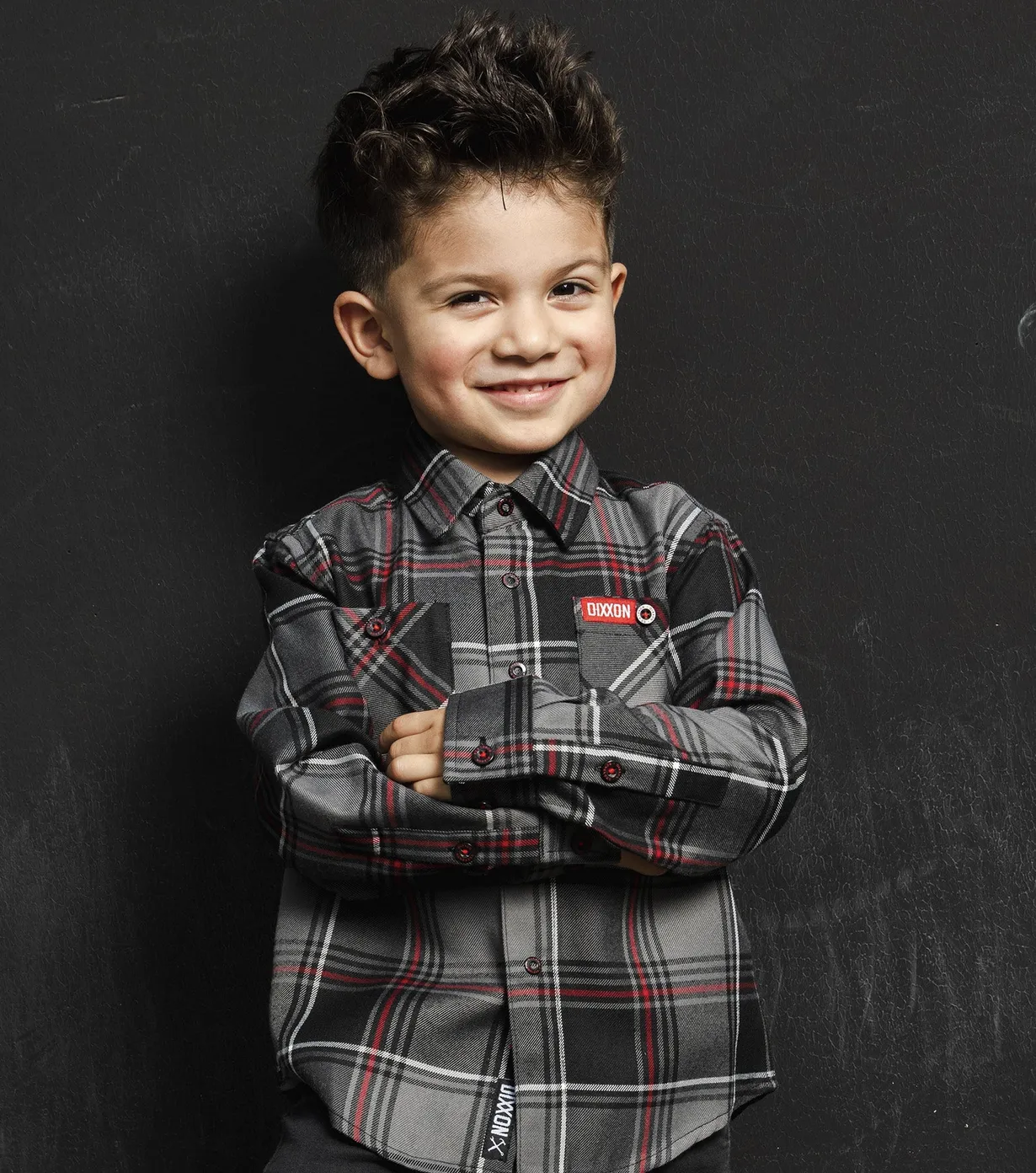 A young boy smiling while wearing a grey, black, and red plaid Dixxon flannel with his arms crossed against a dark textured background. The shirt features a red Dixxon logo tag on the chest pocket and a branded tag at the bottom hem.
