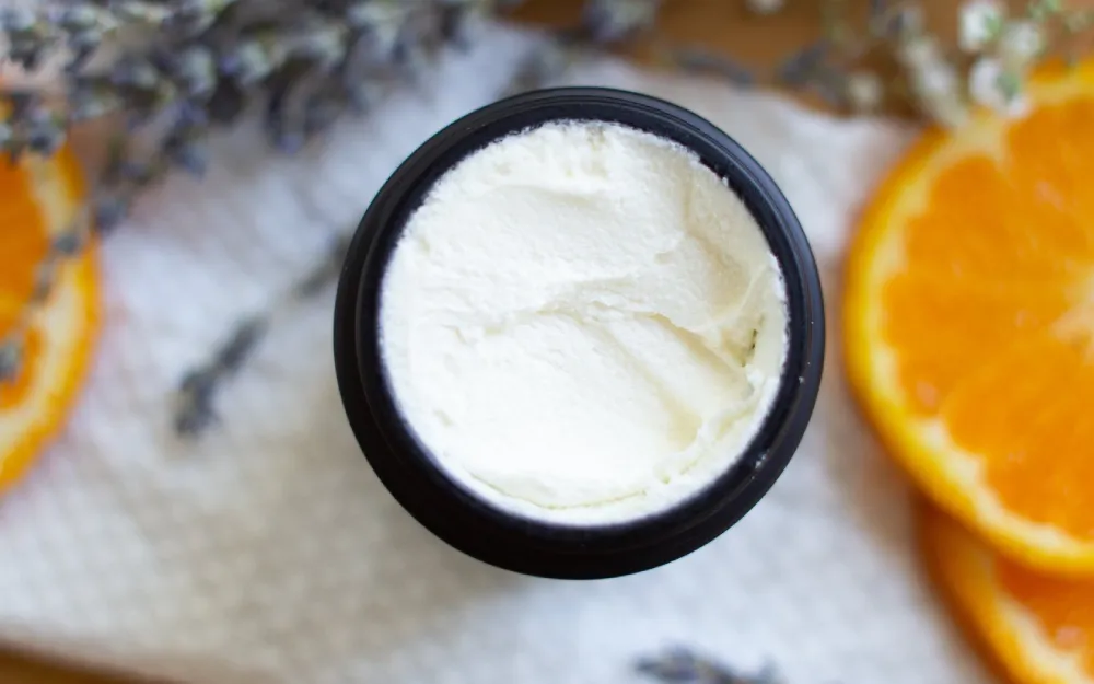 Overhead view of a jar of rich, white cream or balm, possibly a tallow-based product, surrounded by orange slices and lavender, representing natural ingredients for a healthy skincare routine
