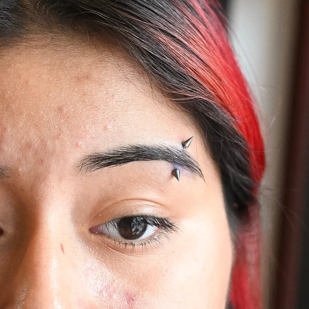 A close-up view of an upper face featuring a vertical eyebrow piercing accented with sharp silver spikes.