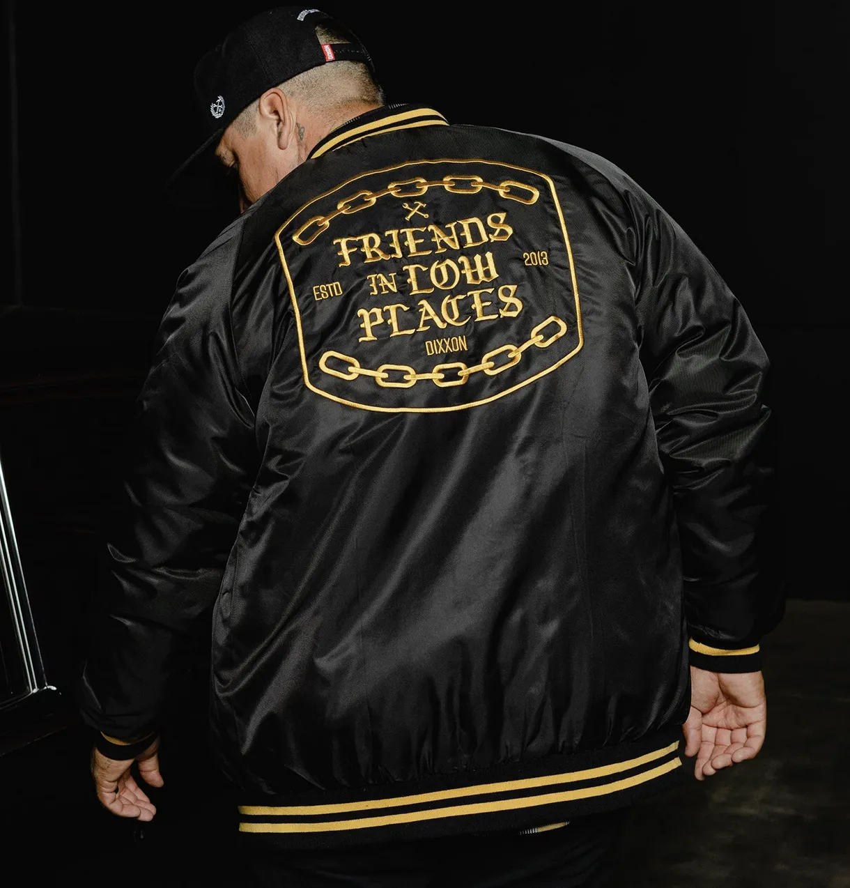 Rear view of a person wearing a black satin bomber jacket featuring a large gold-embroidered patch that reads "Friends in Low Places," "Dixxon," and "ESTD 2013" encircled by a chain design. The person is also wearing a black Dixxon logo hat and the jacket has yellow striped detailing on the cuffs and waistband.