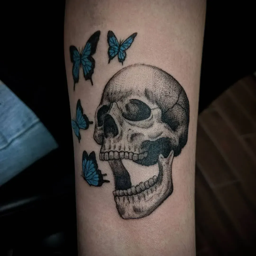 A black and grey stippled skull with an open jaw on a forearm, surrounded by four vibrant blue butterflies.