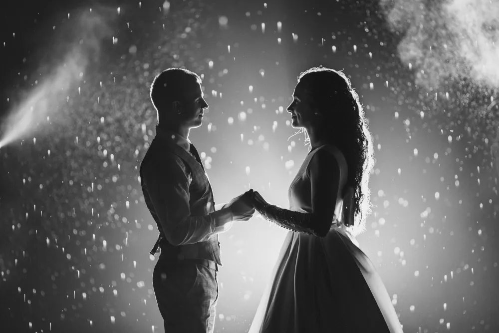 A black and white, dramatic low-light wedding photograph of a bride and groom holding hands and facing each other, backlit by a spotlight, with bright bokeh-like droplets of artificial snow from a snow machine falling around them.