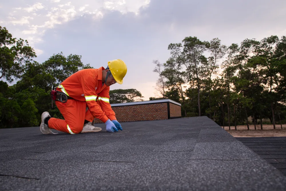 A worker in an orange jumpsuit, yellow hard hat, and blue gloves is kneeling on a dark, flat, granulated roof, inspecting or performing a Roof Repair. The background features trees and a cloudy sunset sky.