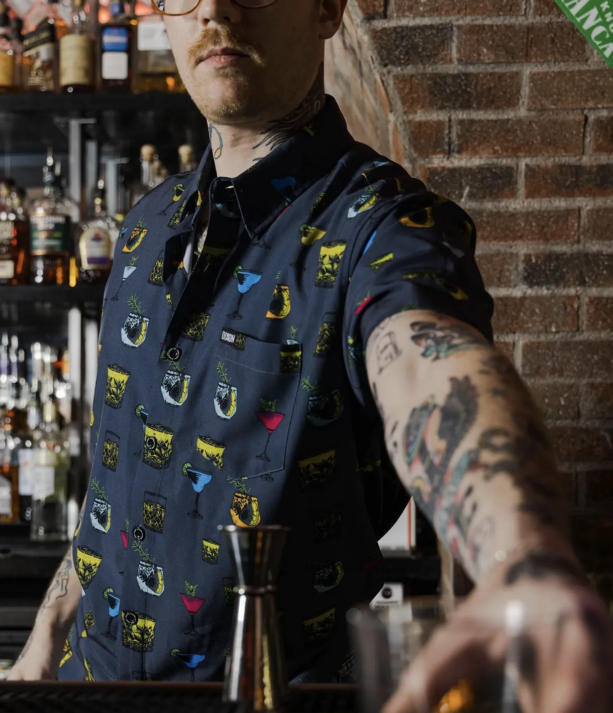A close-up of a tattooed bartender wearing a navy Dixxon Party Shirt featuring a vibrant, repeating pattern of various cocktails, garnishes, and glassware.