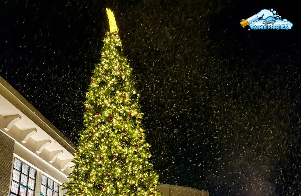 A large, brightly lit outdoor christmas tree with a star topper against a dark night sky, as artificial snow from a snow machine falls around it, creating a festive winter scene next to a building.