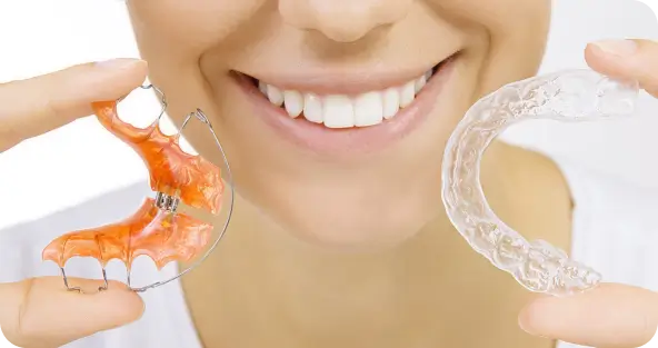 A smiling patient holding a traditional metal Hawley retainer and a clear plastic Essix retainer at Issa Dental Group.