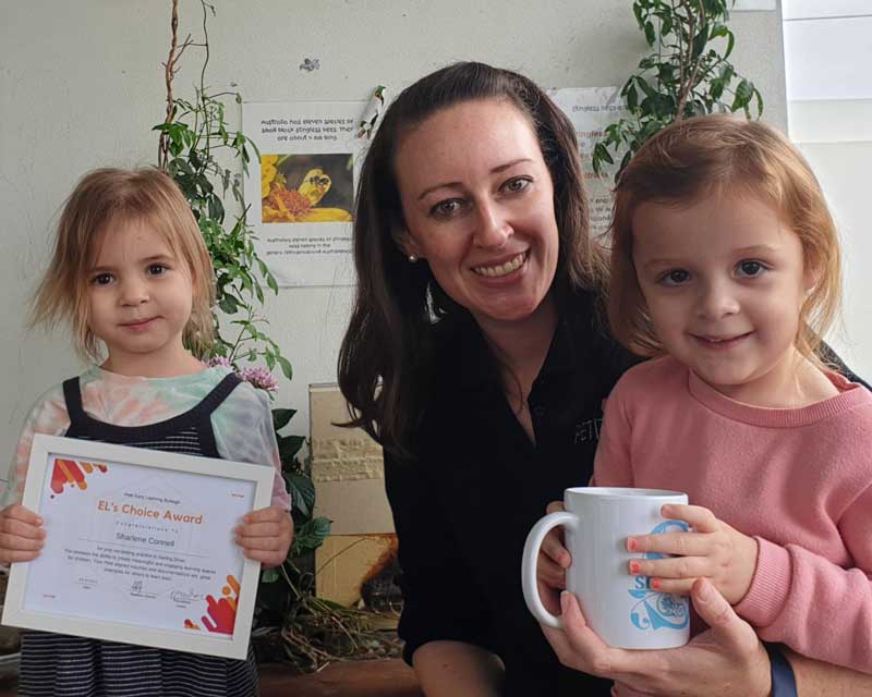 An educator sits between two children. The child on the left holds a framed certificate and the child on the right holds a gift. The educator is receiving an award and gift for Petit ELJ Burleigh's Educational Leader's Choice Award of the month for their personal growth and professional development.