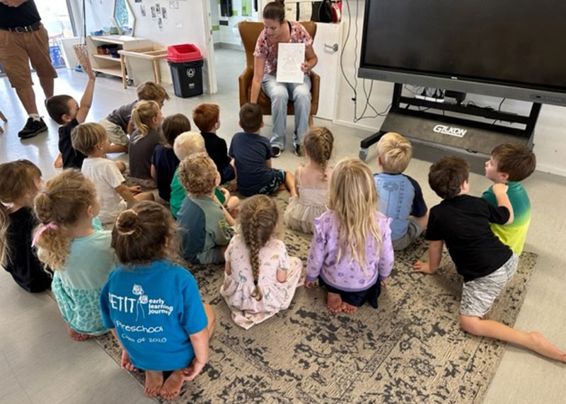 Volcanologist mum sits at the front of the studio holding up a drawing. In front of her preschool children listen as they engage her discussion.