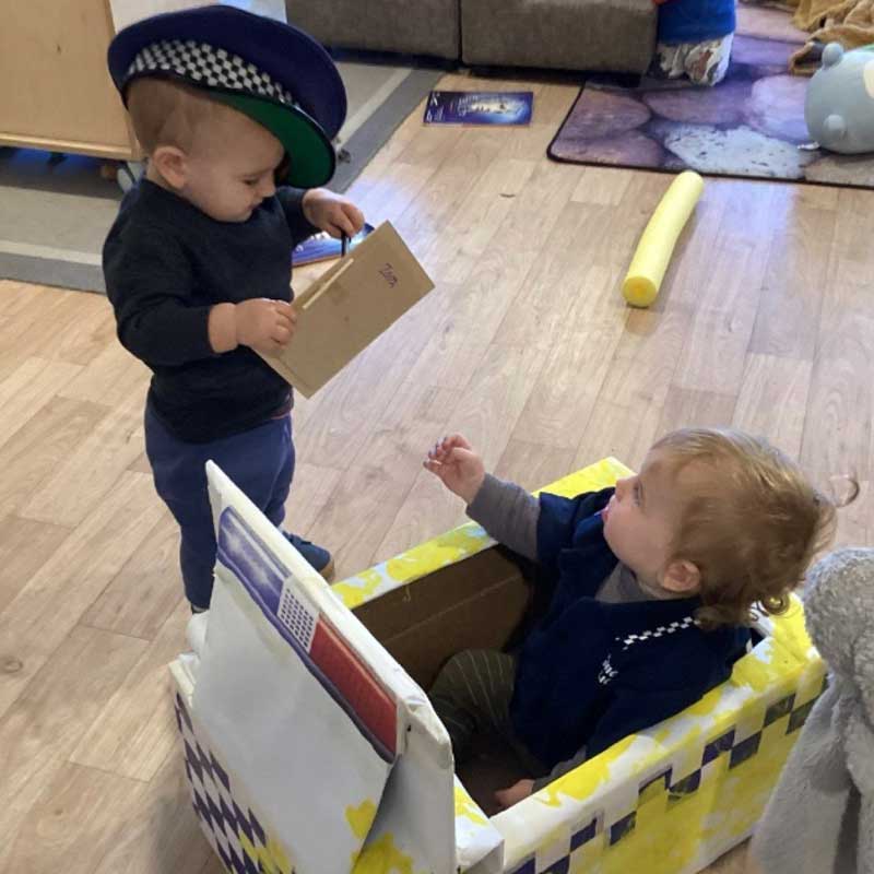 A child wearing a policeman's hat writes a ticket for another child sitting in a police car made from a cardboard box. Both children look engaged in their imaginative play and its numerous benefits.