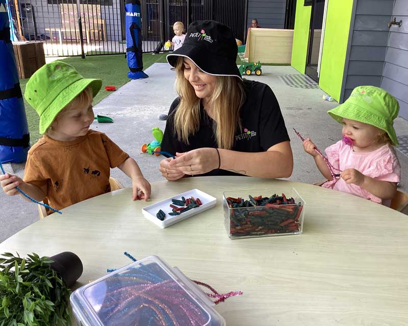 Educator Hannah Sanders from Petit ELJ Pimpama sits between two children in an outdoor space and engages them in loose-parts play. 