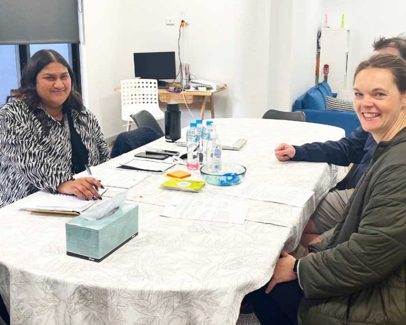 An educator from Petit ELJ Kew sits down at a table to talk about a child's development and learning with a family at a parent-teacher meeting. The educator holds a pen and there are water bottles, a tissue box and forms on the table.