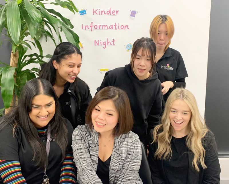 Educators from Petit ELJ Kew gather around a table to review a presentation. They are smiling and laughing. Behind them on the wall is a sign that reads Kinder Information Night. Next to the sign is a green indoor tree.