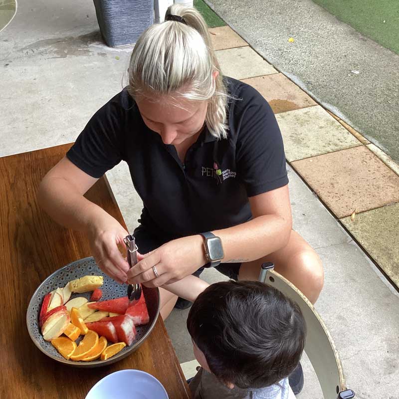 Sammy who believes working with children is a calling, crouches next to a young child and helps them to use a pair of tongs to serve fruit.