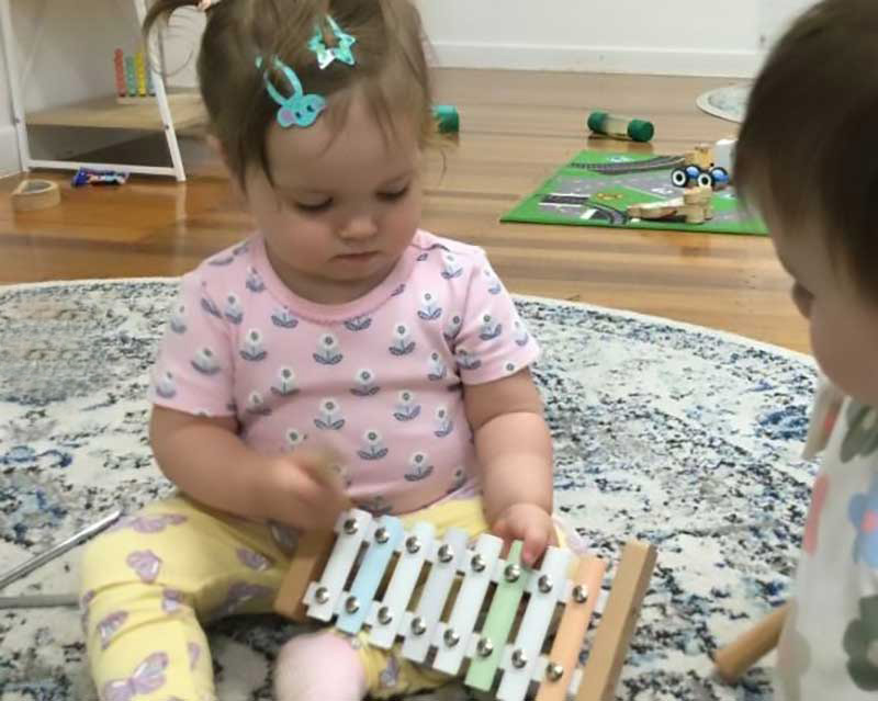 Baby in pink t-shirt and yellow pattered leggings sits on a circular mat holding a musical xylophone in one hand and a blurred mallet in the other. Partially hidden, a second child's face looks on as they play a musical group game.