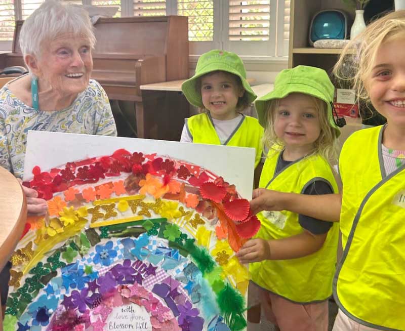 Three children from Petit Early Learning Journey hand a happy Ozcare resident a handmade gift. In the background is a piano and window.\ through which sunlight is shining through.