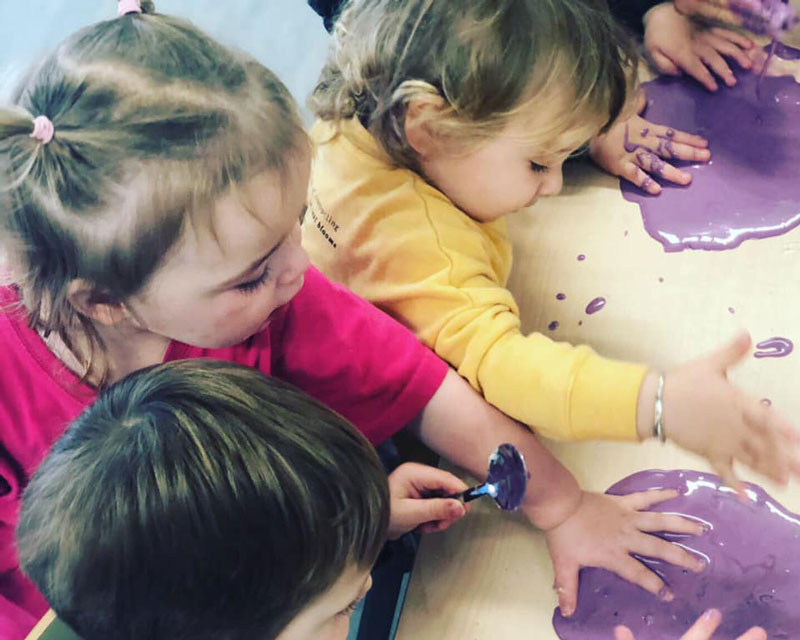 Children crowd around a table to press their hands into an edible slime recipe. One child holds a spoon as they engage in mess play.