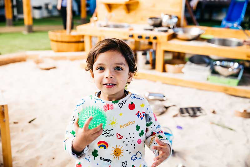 A child in a colourful decorative shirt holds a green spiked ball. He stands in a sandpit and behind him is a pretend kitchen build for imaginative play.