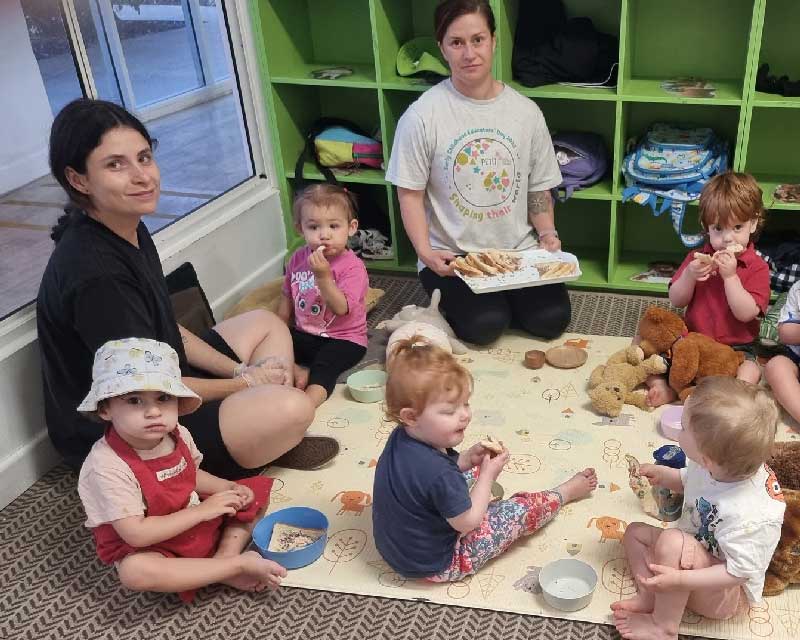 A group of toddlers and two educators sit on a mat with their teddy bears for a teddy bear's picnic.