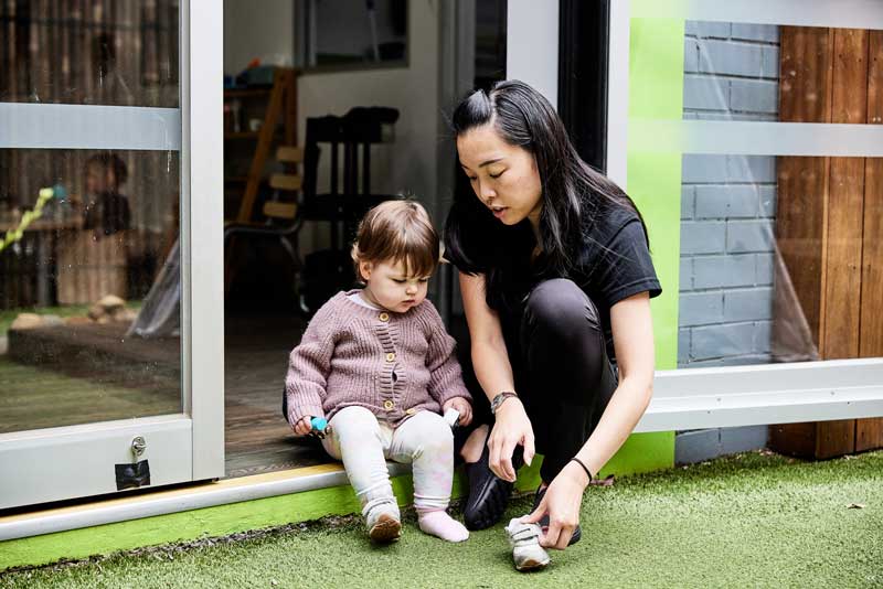 An educator crouches next to a toddler in the doorway between a studio and an outdoor environment helping them with their shoes while listening to their ideas and inspiring them in meaningful conversation.