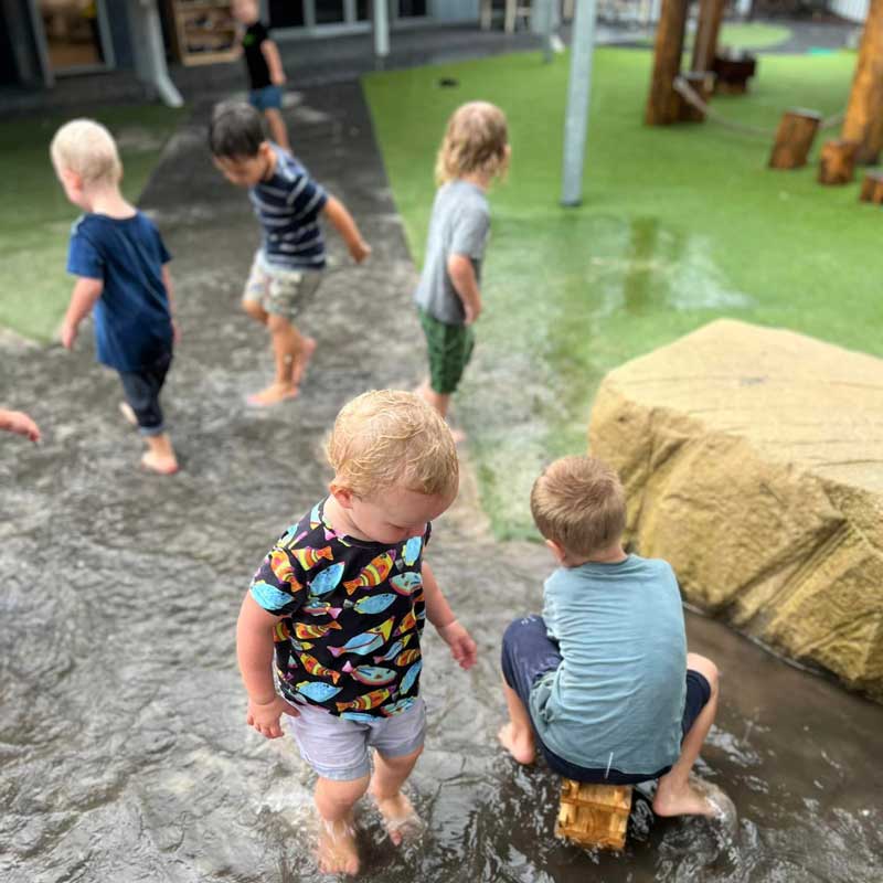 Children splash about in the water having fun with other children as they play. One child sits on a small toy tractor while other children jump about in the puddles. Early Childhood Education and Care centres are one place children can find friends.