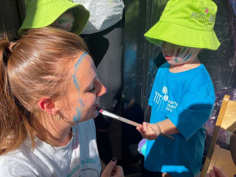 Lead Educator Emma Watkins from Petit ELJ Elderslie interacts in cultural celebrations with a child who paints her face with a pink paintbrush.