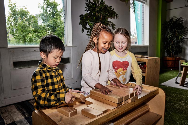 Three children stand behind a bench sorting through different lengths of cut wooden blocks. Wooden blocks are perfect stocking stuffers ideas.
