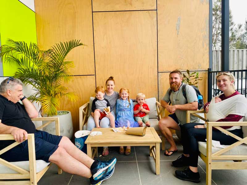 A family sits together on some comfy chairs inside a Petit Early Learning Journey centre where they are involved in their children's early childhood education and enjoying a bite to eat at an end-of-year event.