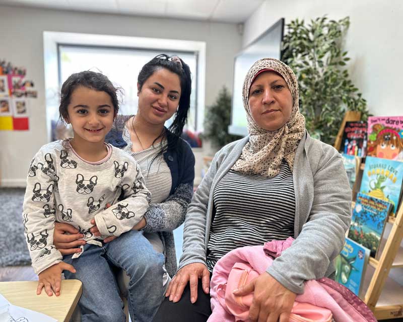 Two women sit in a Petit ELJ studio where parent involvement and partnerships are important. Children's books are on display to their left. A happy child sits on the younger woman's lap.