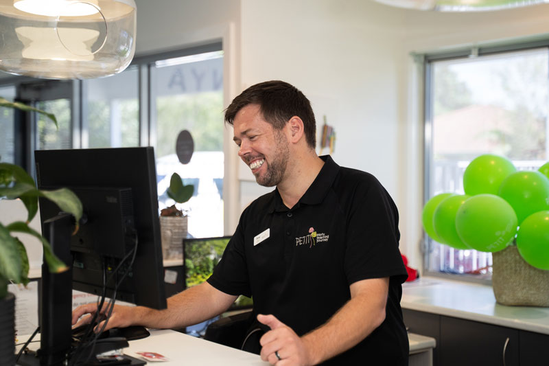 Centre Director Doug at Petit ELJ Caloundra engages with the Petit ELJ Educator Portal to participate and review several professional development examples. Doug stands behind a reception desk with a monitor. A door to his left leads to the carpark and behind him in a basket are several green balloons.