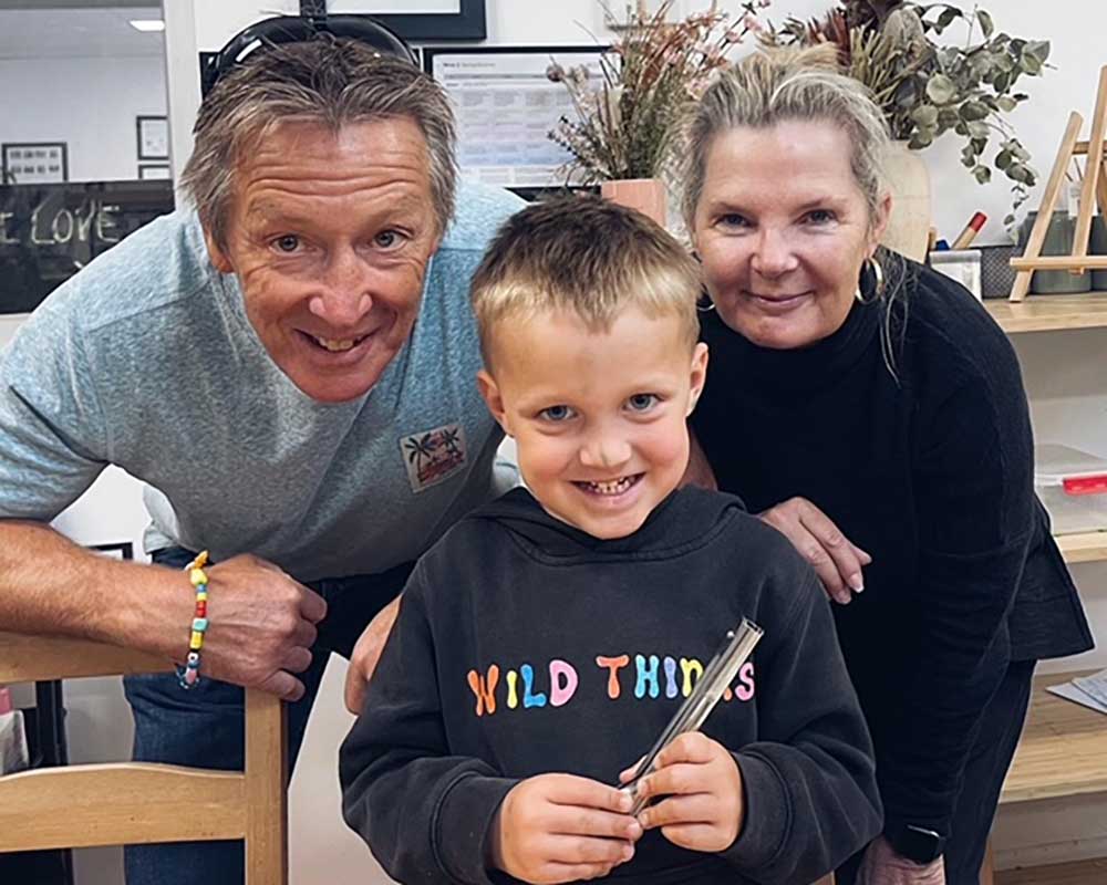 A family gathers together to celebrate in one of the early learning childhood studios with a family picture. A young child stands at the front holding a pair of silver tongs. He grins. His smile is reflected in the faces of his family bending down to look over his shoulder at the camera.