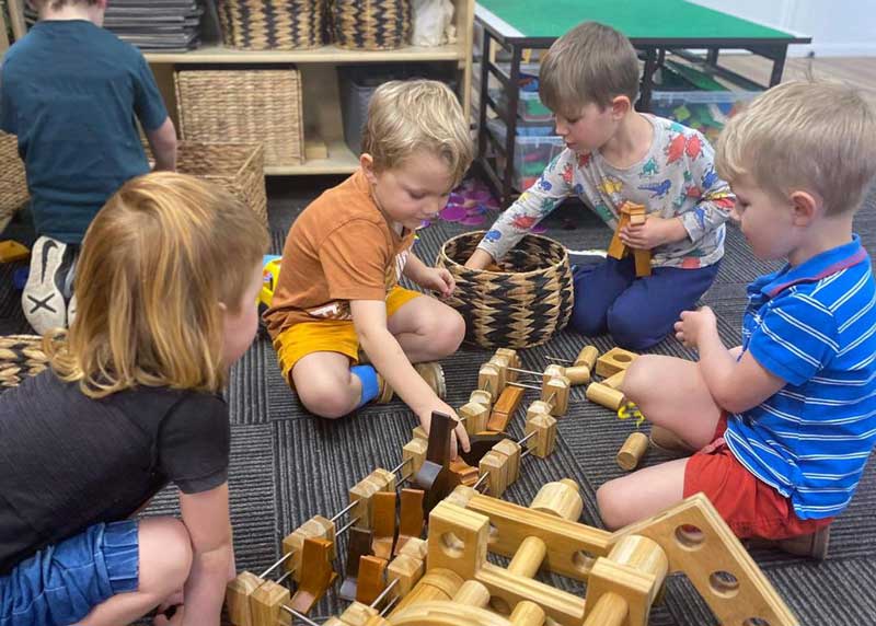 A group of five preschool-aged children sit in a group playing together in an indoor early education and care environment. They are developing children's social skills as they assemble a wooden loose parts project.