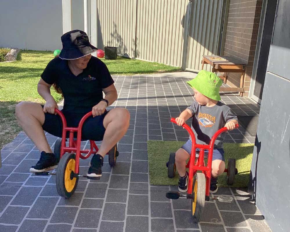 An educator rides a red tricycle next to a young child on a similar vehicle in an outdoor environment at Petit Early Learning Journey Coffs Harbour, the 2023 Perpetual Award winner for Centre of the Year. The educator is smiling, having fun and encouraging the child to pedal as he looks at the educator's feet.