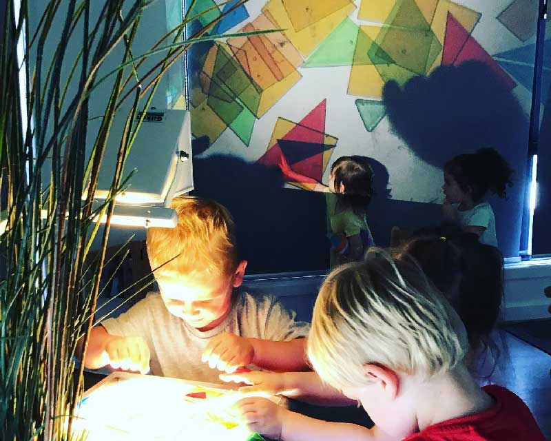 Children engage in a light and shadow play experience as they surround a light projector manipulating shapes that reflect upon a wall.