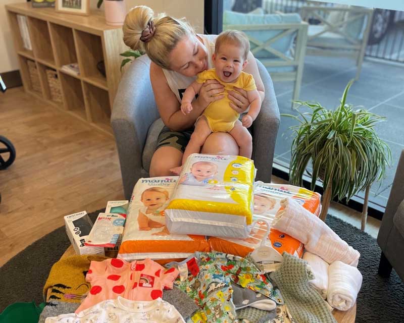 Parent sits holding a baby behind a table filled with family donations of nappies and clothes for Hearts of Purple in 2022.