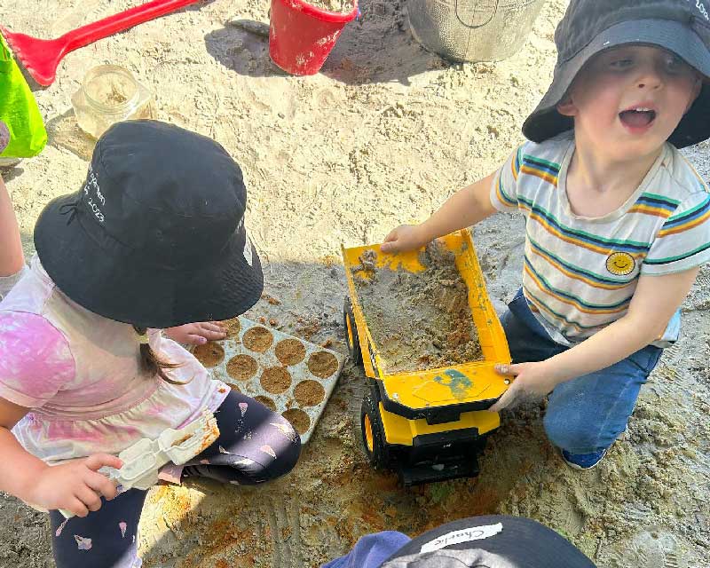 Two children sit in a sandpit. One child in a horizontally colourful stripped shirt plays with a truck. The back of the truck is filled with wet sand. The other child in a pink dress fills a baking tray with damp sand. Behind them is a red and a silver bucket, and a red plastic rake.